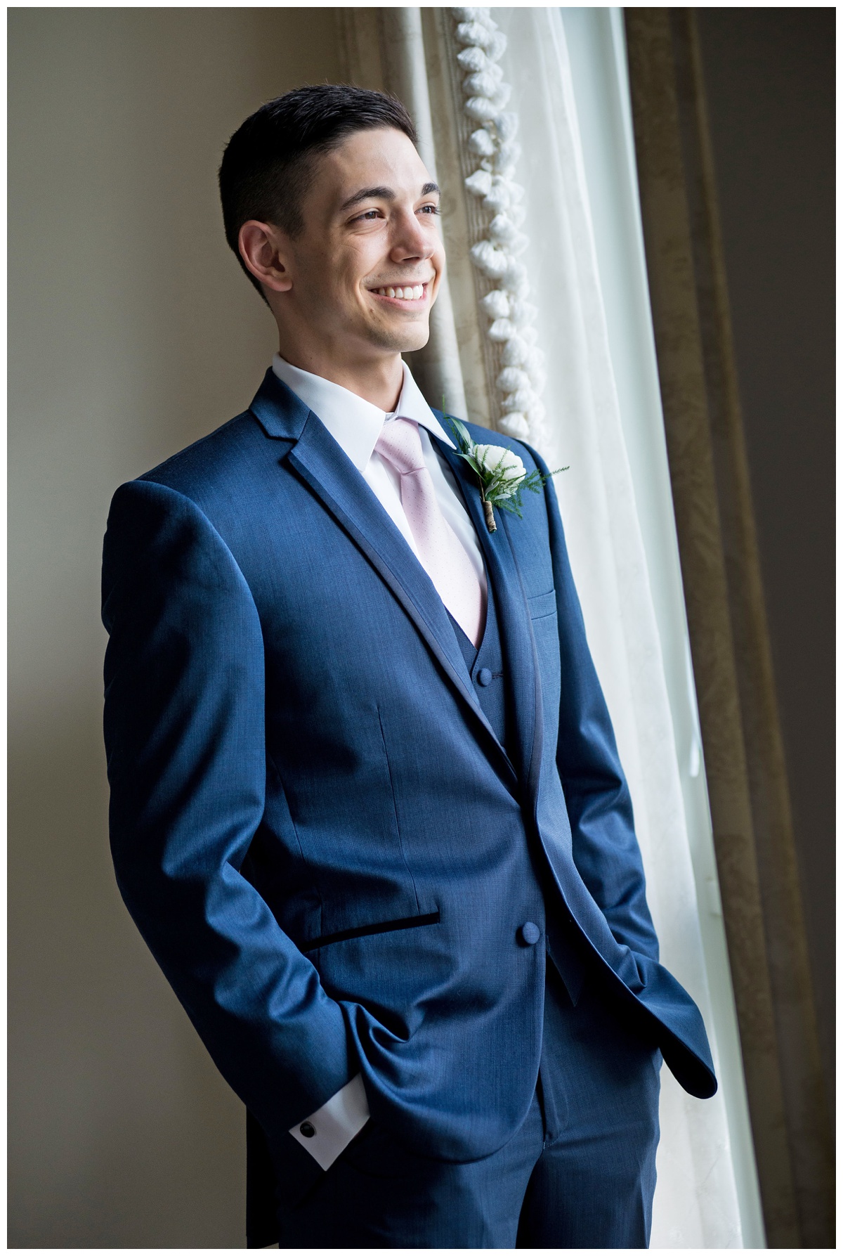 photo of smiling groom at june wedding in the summer - looking out window in blue suit with light pink tie at The Oaks Waterfront Inn - getting ready photo