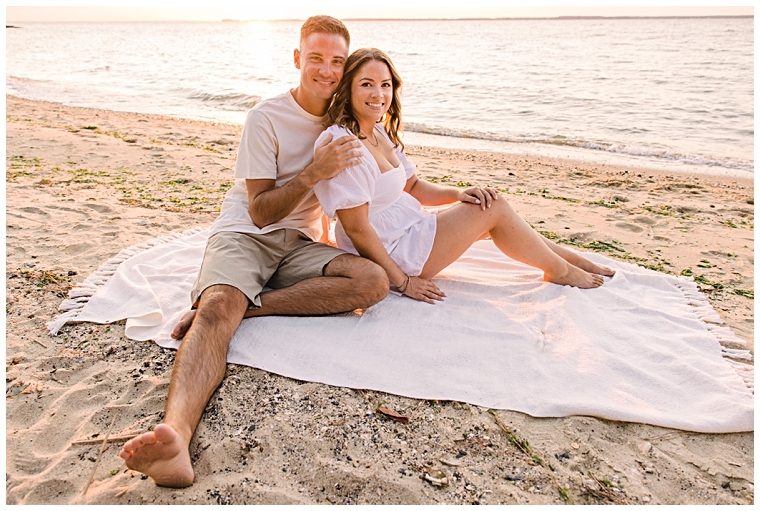 The newly betrothed couple sits on a blanket in the sand enjoying the waterfront