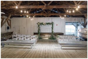 A beautiful ceremony site in the Boat House at The Chesapeake Bay Maritime Museum is decorated with greenery and white florals by Seaberry Farm