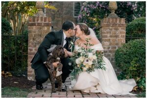 The bride and groom share a kiss as their puppy poses for a portrait