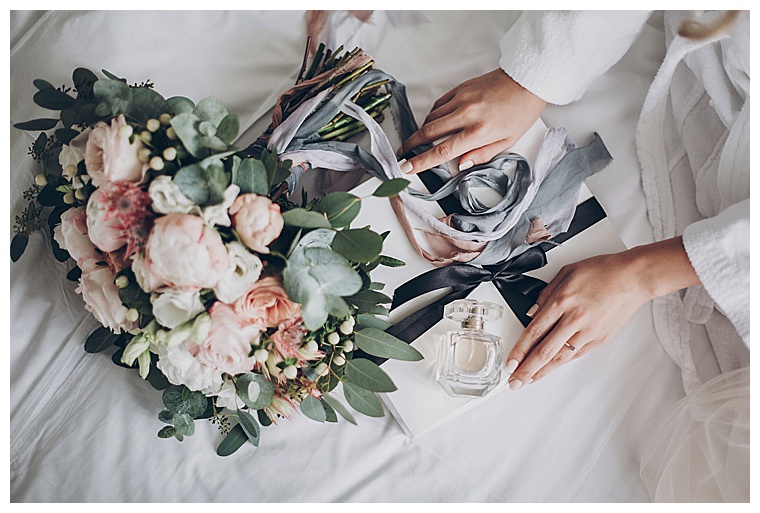 Modern wedding bouquet, perfume bottle, and gift box on white bed. Bride holding white box for morning boudoir before wedding ceremony.