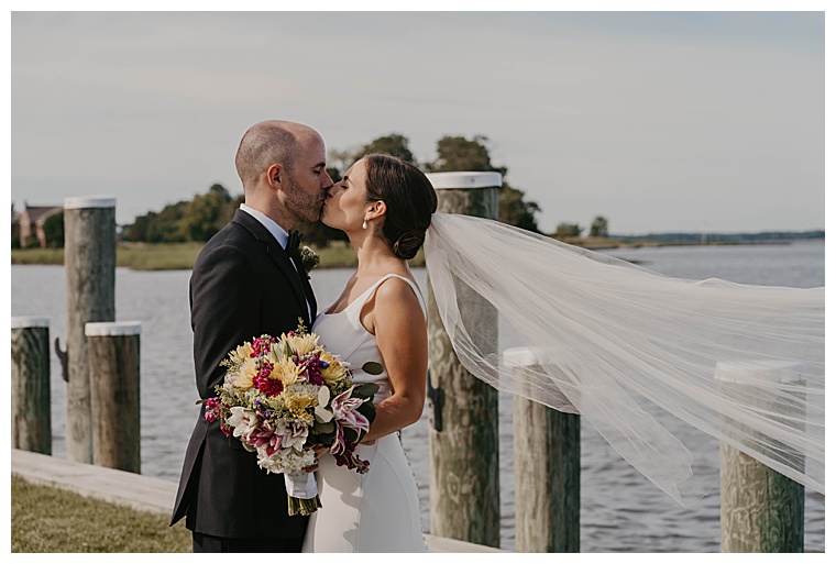 The bride and groom embrace in a kiss at the waterfront at the Chesapeake Bay Maritime Museum and the bride's veil flows in the breeze