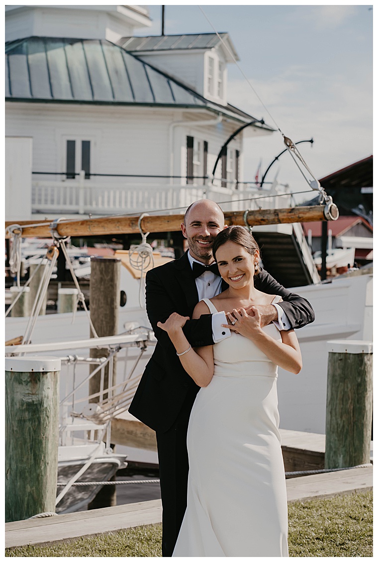 The groom hugs his new wife from behind with the beautiful Chesapeake Bay Maritime Museum lighthouse in the background
