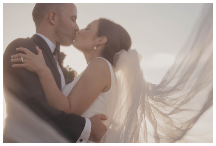 The bride's veil flows in the wind as she kisses her new husband