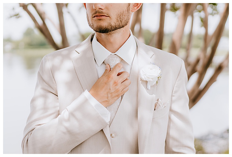 The groom adjusts his tie as he awaits his first look with his soon to be wife. He is looking sharp in a sand colored suit and a white rose boutonniere. 