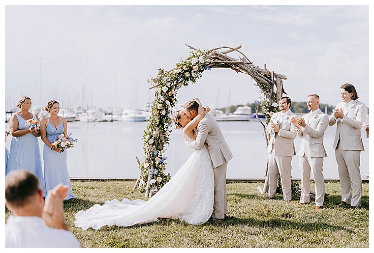On the waterfront at the Inn at Haven Harbour, under a custom made driftwood arch with roses and greenery, the newlyweds embrace in their first kiss as husband and wife as their bridal party cheers them on