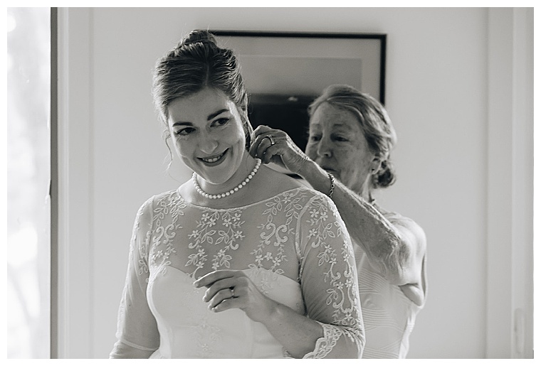 The mother of the bride helps the bride with her pearl necklace as they get ready for the ceremony