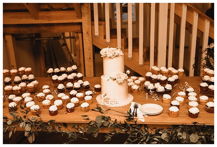 A stunning and sleek two tiered wedding cake is surrounded by a sea of wedding cupcakes in a variety of delicious flavors on this dessert bar