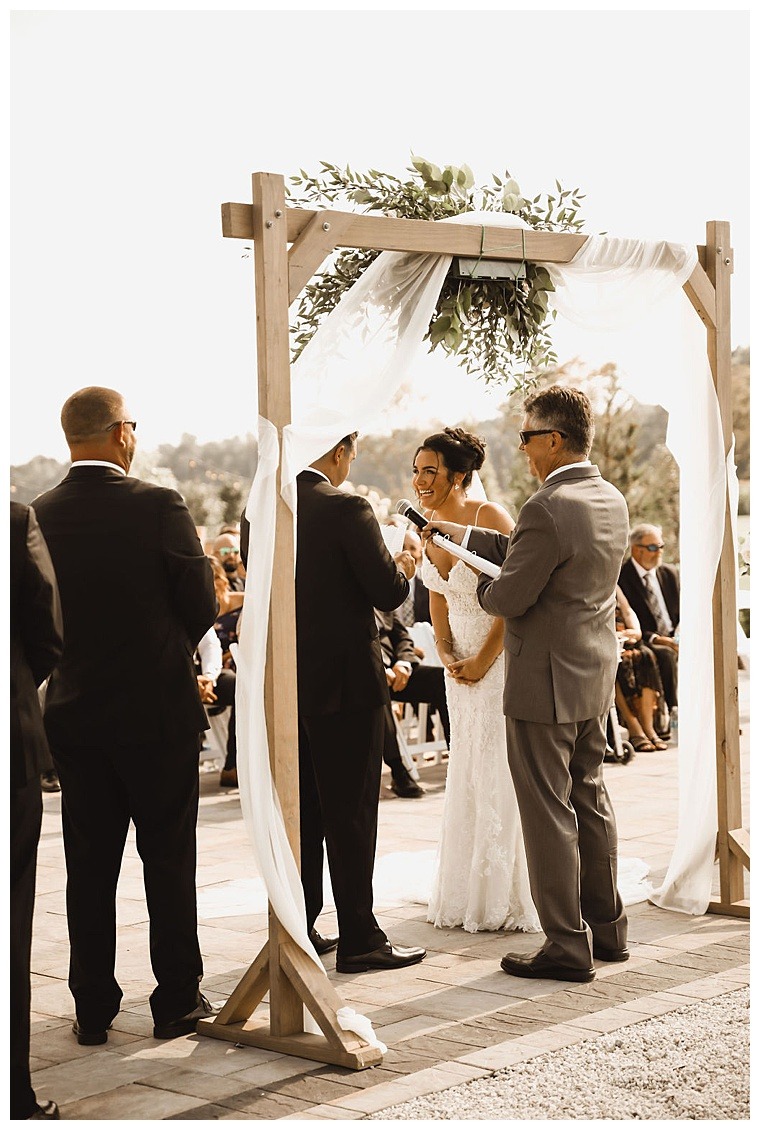 The bride and groom exchange their wedding vows under a wooden archway decorated with greenery and white linens at Breckenridge Barn