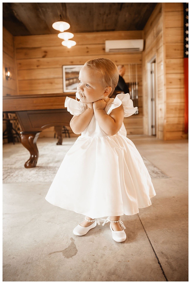 The flower girl awaits her big moment at Breckenridge Barn wearing a simple but elegant white flower girl dress with pearly white ballet flats