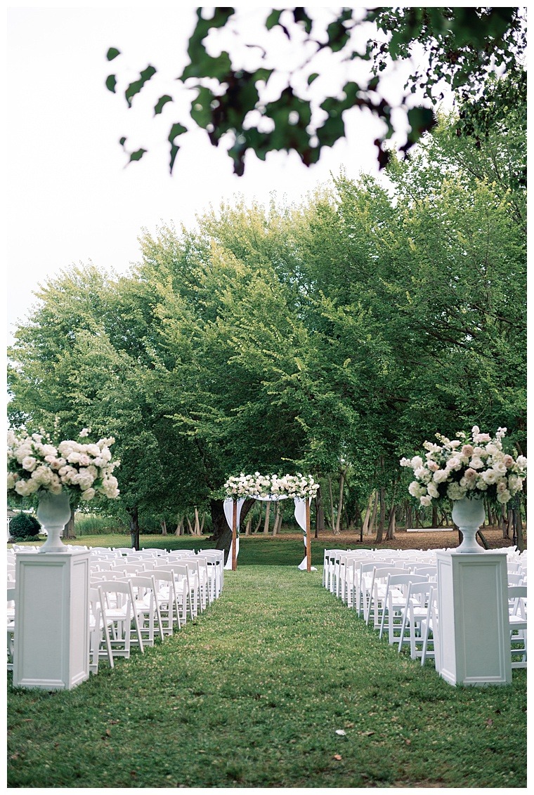 A white wedding ceremony is framed in greenery from the lush trees to the white rose bouquets lining the aisle and the archway
