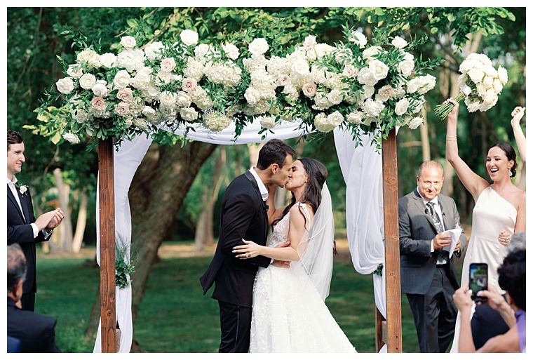 The bride and groom embrace in their first kiss as husband and wife under a stunning archway decorated in white roses and greenery as guests cheer and applaud