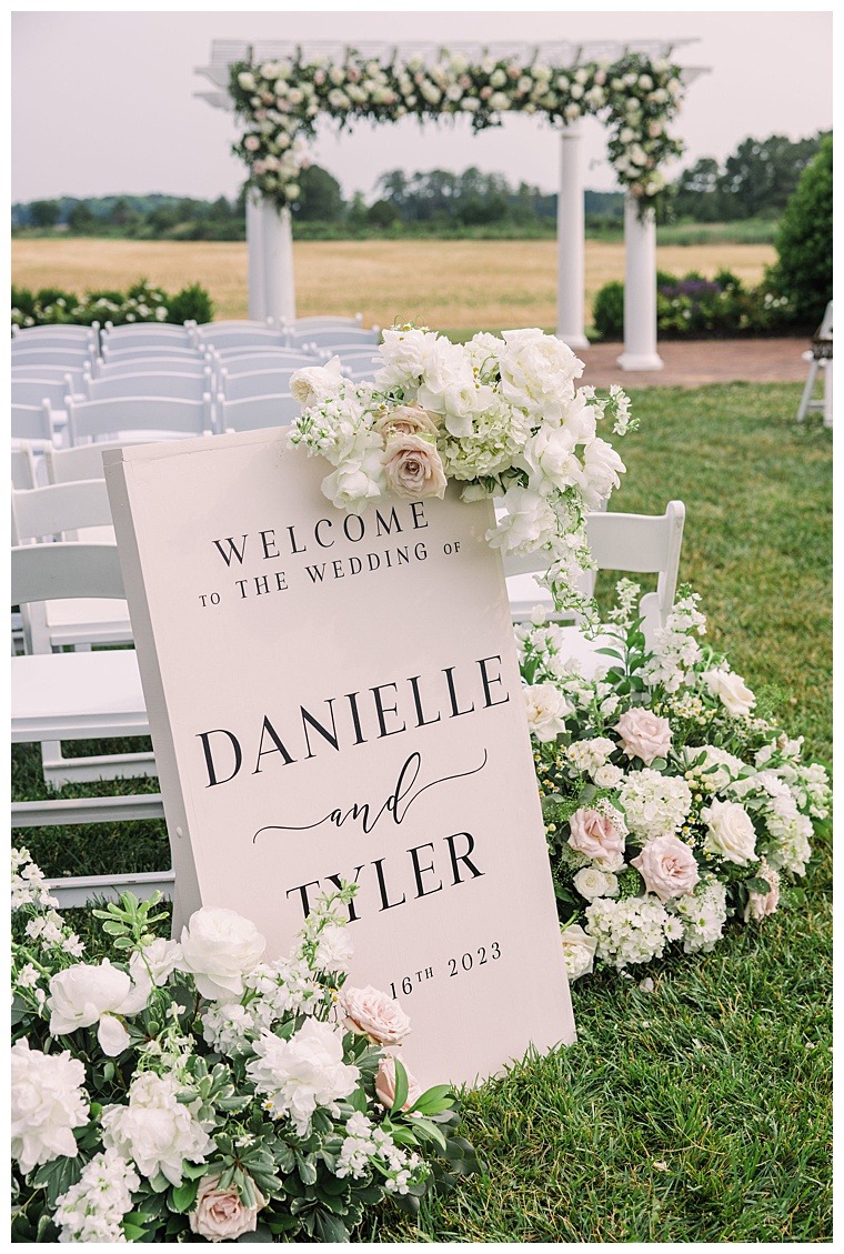 A custom welcome sign to greet guests as they arrive at Danielle and Tylers ceremony site adorned with beautiful florals and greenery to match the archway where they newlyweds will exchange their vows