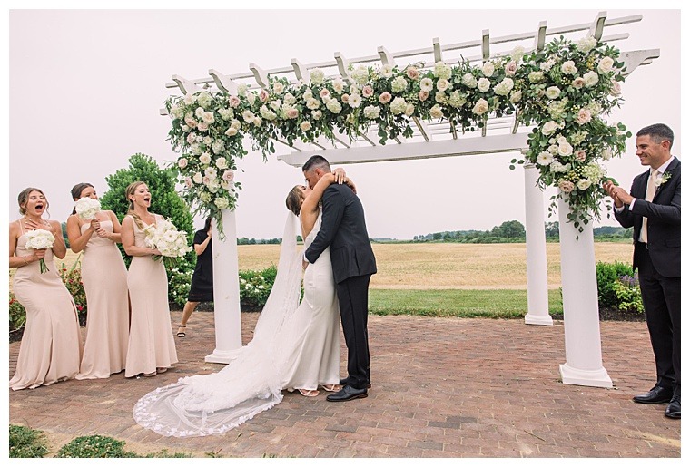 The bride and groom share their first kiss as husband and wife under a truly stunning archway decorated with white roses and lush greenery as their bridal party cheers for the newlyweds