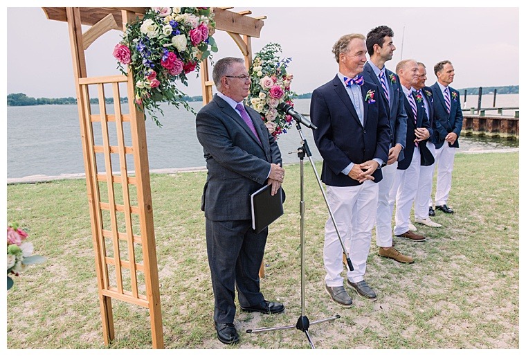 The groom and his groomsmen, dressed in white pants and blue blazers, await the bride as she processes down the grassy aisle to her waterfront ceremony