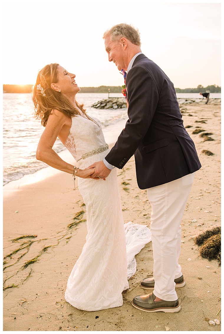 The bride and groom, in a classic white gown and white pants and sperrys, dance on the beach in the sunset over the Tred Avon River